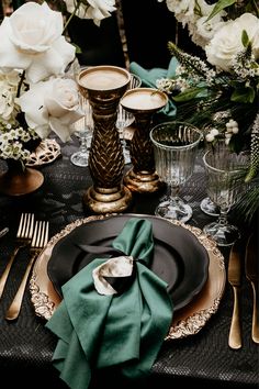 a black and gold place setting with green napkins, silverware and white flowers