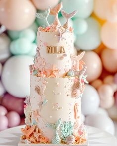 a three tiered cake decorated with sea shells and starfish on a table in front of balloons