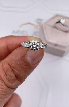 a person holding an engagement ring with three stones on it and a box in the background