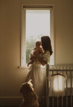 a woman holding a baby standing next to a crib in front of a window