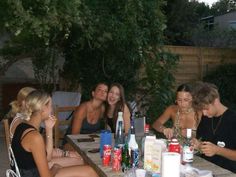 a group of people sitting around a wooden table with bottles and drinks in front of them