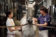 a man and woman standing next to a white horse in a stable with their hands on the bridle