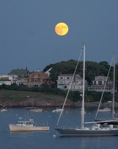 a full moon is seen over boats in the water near houses and trees on land