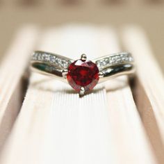 a red heart shaped diamond ring sitting on top of a wooden table