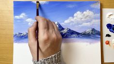 a person is holding a paintbrush and painting on a piece of paper with mountains in the background