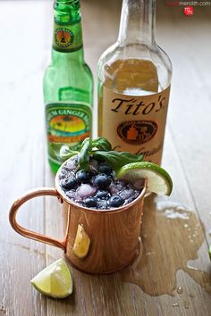 a copper mug filled with blueberries and limes next to two bottles of booze