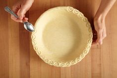 a person holding a spoon over a pie crust on a wooden table with a knife and fork