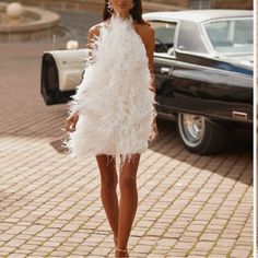 a woman is walking down the street wearing a white dress with feathers on it and an old car in the background