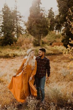 a man and woman are walking through the grass in an open field with tall trees