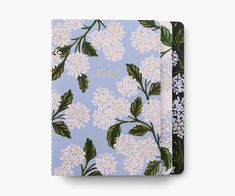 a blue notebook with white flowers and green leaves on the cover, next to a black pen