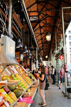 a woman standing in front of a market filled with fruits and vegtables