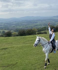 a man riding on the back of a white horse across a lush green field with mountains in the background