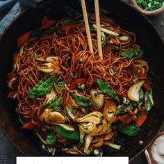 a wok filled with noodles and vegetables next to chopsticks on a table