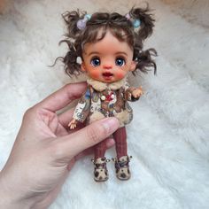 a hand holding a small doll on top of a white fur covered floor next to a stuffed animal