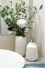 a white vase with flowers in it sitting on a bathroom counter next to a sink