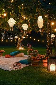 an outdoor seating area with lanterns and pillows on the grass, surrounded by flowers and trees