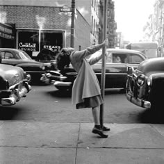 a black and white photo of a woman leaning on a pole in front of parked cars