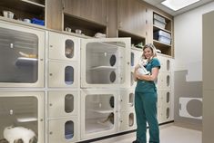 a woman in scrubs holding a white cat and looking at the camera while standing next to cabinets