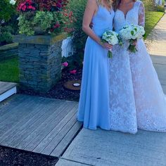 two beautiful women standing next to each other in front of some flowers and bushes on the sidewalk