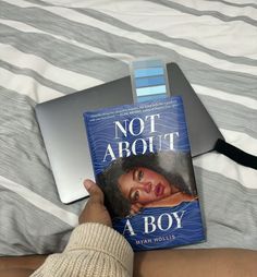 a person holding a book in their hand on top of a bed next to a laptop