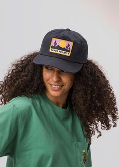 a woman with curly hair wearing a green shirt and black hat, smiling at the camera