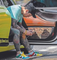 a man with tattoos sitting on the hood of a car looking at his cell phone