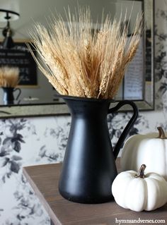 a black vase filled with dry grass next to two white pumpkins on a table