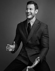 a black and white photo of a smiling man in a suit sitting on a chair