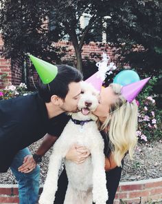 a man kissing a woman while holding a dog wearing party hats on their heads and nose