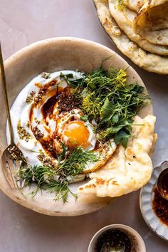 an egg and some herbs in a bowl on a table next to pita bread