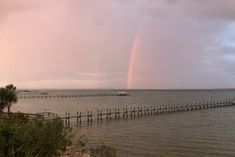 a rainbow appears over the ocean with a long wooden pier in the foreground and a boat out on the water