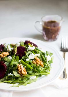 a white plate topped with salad next to a fork and glass filled with dressing on top of a table