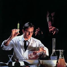 a man in white shirt and tie holding a lit candle over a table filled with laboratory equipment