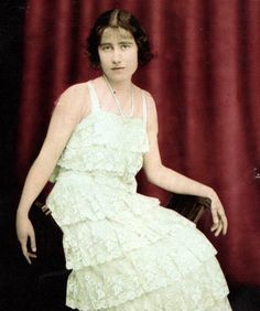 an old photo of a woman in a white dress sitting on a chair with a red curtain behind her