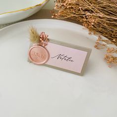 a white plate topped with a pink place card and dried flowers next to a vase filled with dry grass