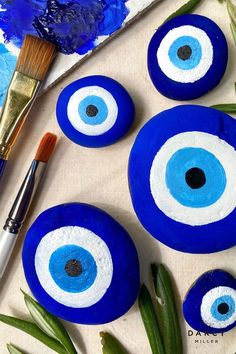 three blue evil eyes painted on rocks next to green plants and paintbrush with gold leafy stems