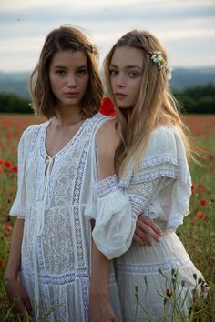 two young women standing in a field with red flowers on their head and one wearing a white dress