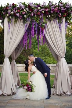 a bride and groom kissing in front of an outdoor wedding ceremony arch with purple flowers