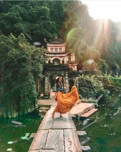 a woman in an orange dress is walking on a bridge over water with trees and bushes