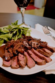 a white plate topped with meat and greens next to a glass of wine on top of a table