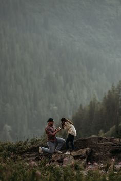 a man and woman standing on top of a rocky hill next to trees in the distance