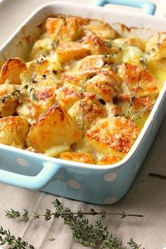 a casserole dish with potatoes and meat in it sitting on a table next to some herbs