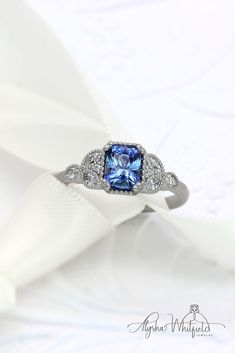 a ring with a blue stone surrounded by diamonds on a white satin background, in the shape of a heart