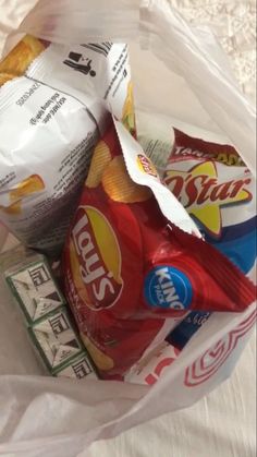 a plastic bag filled with chips and snacks on top of a white tablecloth covered bed