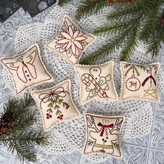 four embroidered christmas ornaments on a doily with pine cones and evergreen branches in the background