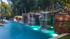 an outdoor swimming pool with waterfall and artificial lighting in the water, surrounded by lush greenery