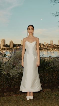 a woman in a white wedding dress standing on the grass near some bushes and trees