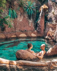 two people are sitting on rocks in the water near a rock pool and some trees