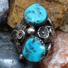 This is an Old Navajo Double Turquoise Ring Intricate Appliques Size 7.25 Sterling Silver C1970s. This size 7.25 Native American ring includes two polished turquoise nugget stones (one with tiny bits of pyrite) surrounded by intricate twisted wire appliques, flowers, feathers and raindrops. The ring tests positive for sterling silver. The face measures 1 inch and wraps around to the sides. The ring weighs 9.1 grams.  2235635 Western Style Turquoise Ring Collectible, Vintage Concho Ring Jewelry, Vintage Adjustable Concho Rings, Vintage Turquoise Multi-stone Rings, Vintage Multi-stone Turquoise Jewelry, Vintage Turquoise Multi-stone Jewelry, Vintage Turquoise Concho Ring, Vintage Turquoise Ring With Inlay For Anniversary, Vintage Blue Turquoise Ring For Anniversary