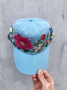 a hand holding up a blue hat with floral embroidery on the front and side, against a gray wall